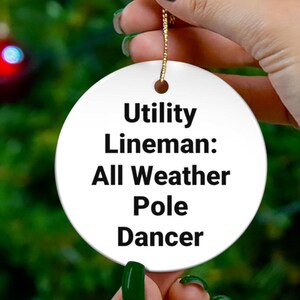 May include: A round white ceramic ornament featuring the black printed text Utility Lineman: All Weather Pole Dancer. The ornament has a small hole at the top with a thin gold string attached for hanging, presenting a smooth and glossy surface finish.