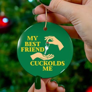 May include: A round, green ceramic ornament with the text "MY BEST FRIEND CUCKOLDS ME" in yellow. The ornament features a graphic of two hands exchanging keys. A gold string is attached for hanging.