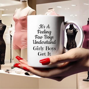 Può includere: Una tazza di ceramica bianca con la scritta "It's A Feeling Few Boys Understand Girly Boys Get It" in corsivo nero. La tazza è tenuta in mano con lo smalto rosso. Lo sfondo include manichini che indossano lingerie.
