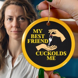May include: A round, black and yellow keychain with the text "MY BEST FRIEND CUCKOLDS ME". The keychain features illustrations of hands holding a key. The background includes a person.