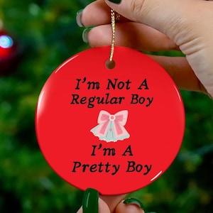 以下が含まれることがあります： 「I'm Not A Regular Boy, I'm A Pretty Boy」の黒い文字が入った、丸い赤いクリスマスオーナメント。ピンクと青のリボンのグラフィックが最初の行の下に配置されています。オーナメントには、吊り下げ用の金色の紐が付いています。
