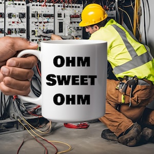 May include: White ceramic mug with the text "OHM SWEET OHM" in bold black letters. The mug is held in front of an electrician working on electrical panels, wearing a yellow hard hat and safety vest.
