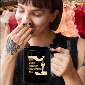 May include: Black ceramic mug with the text "MY BEST FRIEND CUCKOLDS ME" in yellow. The design includes a key and hands. The mug is held by a person with black nail polish and a tattoo on their arm.
