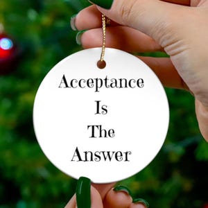 May include: A white, round ceramic ornament with the words "Acceptance Is The Answer" in black script. The ornament is suspended by a gold string, and is held by a hand with green nail polish. The background is a blurred green.