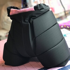 May include: Black padded shorts with a pink sparkly waistband. The shorts have multiple padded sections for protection.