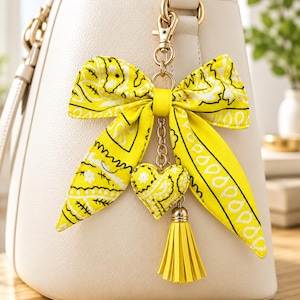 Puede incluir: Un llavero de lazo de tela amarillo brillante con un patrón de cachemira negro, unido a un mosquetón de metal dorado. El accesorio incluye un pequeño dije de corazón acolchado con un estampado de cachemira amarillo y negro y una borla de cuero amarilla.