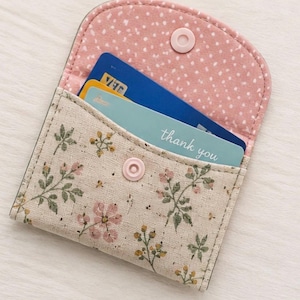 Puede incluir: Un tarjetero de tela con exterior beige y estampado floral de flores rosas y hojas verdes. El interior está forrado con tela rosa de lunares blancos. Un botón a presión rosa claro asegura la solapa. Hay dos tarjetas insertadas dentro.