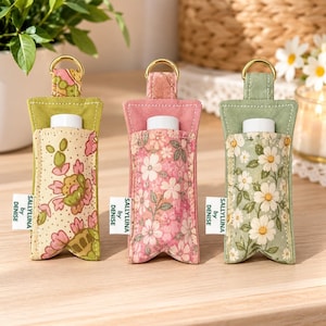 May include: Three fabric lip balm holders with gold-tone metal rings. Each holder features a floral pattern in green, pink, or white and includes a white label reading SALLYLUNA by DENISE. The holders are designed to store a standard lip balm tube.