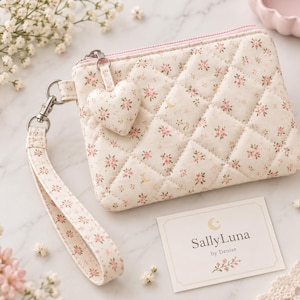 May include: A quilted cream-colored zippered pouch featuring a small pink and green floral pattern. The pouch includes a matching wrist strap and a heart-shaped charm attached to the zipper pull. A white card displays the text SallyLuna by Denise.
