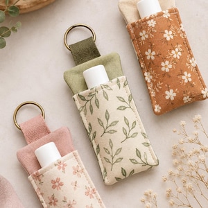 May include: Three fabric lip balm holders with metal key rings. One is cream with pink flowers, one is cream with green leaves, and one is rust orange with white flowers. Each holder contains a white plastic lip balm tube inside a fabric sleeve.