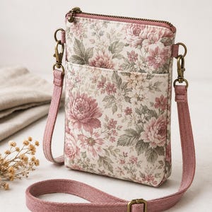 Könnte beinhalten: Eine rechteckige Umhängetasche mit Blumenmuster in Altrosa, Creme und Salbeigrün. Die Tasche verfügt über einen Reißverschluss an der Oberseite, ein offenes Außenfach und einen verstellbaren Schulterriemen aus altrosa Stoff mit Messing.