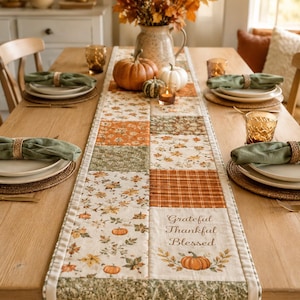 Könnte beinhalten: Ein rechteckiger Tischläufer aus Stoff mit einem Patchwork-Design aus herbstlichen Mustern, darunter orangefarbene Kürbisse, Herbstblätter und Karomuster. Ein Abschnitt zeigt den Text Grateful Thankful Blessed über einer Kürbisgrafik.