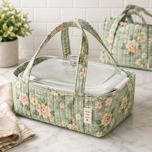 May include: A rectangular quilted casserole carrier in a sage green fabric with a peach and white floral pattern. It features two fabric handles and a small white tag that reads made with love. A clear glass baking dish with a lid sits inside.