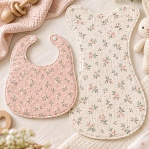 Può includere: Una bavaglino per neonati e una coperta per neonati con un motivo floreale abbinato. La bavaglino è rosa con fiori bianchi e foglie verdi, mentre la coperta è color crema con la stessa stampa floreale. Entrambi hanno una finitura in tessuto.