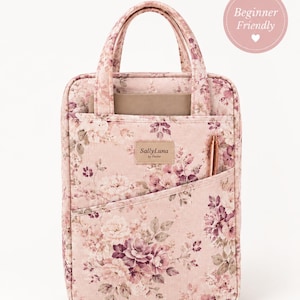 May include: A rectangular fabric tote bag featuring a pink floral pattern with shades of mauve and cream. The bag has two top handles, a front slip pocket containing a metallic pen, and a central label reading SallyLuna by Denise.