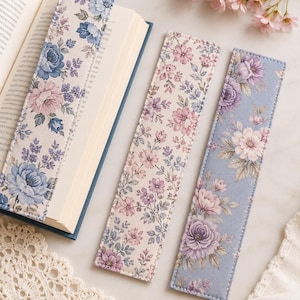 May include: Three rectangular fabric bookmarks featuring floral patterns in shades of blue, purple, and cream. One bookmark is placed inside an open book, while two others lie flat, showcasing detailed botanical prints with roses and small leaves.