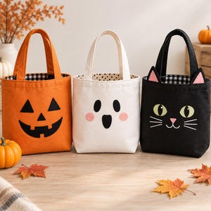 Puede incluir: Tres bolsas de tela con asas y diseños de Halloween: una bolsa naranja con cara de calabaza negra, una bolsa blanca con cara de fantasma y mejillas rosadas, y una bolsa negra con cara de gato, ojos verdes y orejas rosadas.