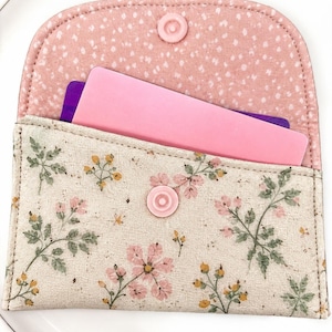 May include: A fabric card holder featuring a beige exterior with a floral pattern of pink flowers, green leaves, and yellow buds. The interior flap is pink with white speckles. A pink plastic snap closure secures the pouch, which contains a pink card.