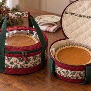May include: Two round, quilted pie carriers featuring a festive holly and berry print on a cream background with solid red quilted borders and dark green fabric handles. One carrier is closed with a clear plastic top, while the other is open.