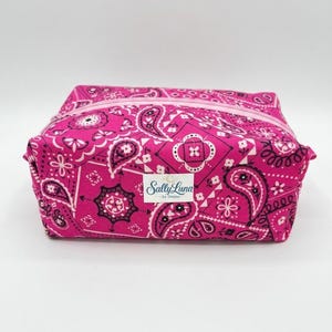 May include: A rectangular, hot pink cosmetic bag with a paisley pattern in black and white. The bag has a matching pink zipper and the text "Sally Luna by Denise" on a white label.