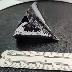Puede incluir: Objeto triangular oscuro y reflectante con una superficie texturizada, posiblemente un cristal o un trozo de silicio. Una regla con marcas en pulgadas se coloca junto al objeto. El objeto mide aproximadamente 5 cm de largo.