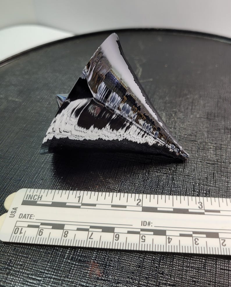 Puede incluir: Objeto triangular oscuro y reflectante con una superficie texturizada, posiblemente un cristal o un trozo de silicio. Una regla con marcas en pulgadas se coloca junto al objeto. El objeto mide aproximadamente 5 cm de largo.