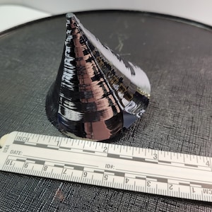 Puede incluir: Un objeto oscuro y reflectante en forma de cono con una base plana, posiblemente una oblea de semiconductor. Una regla blanca con marcas en cm est&aacute; en primer plano. La regla muestra medidas de hasta 10 cm.