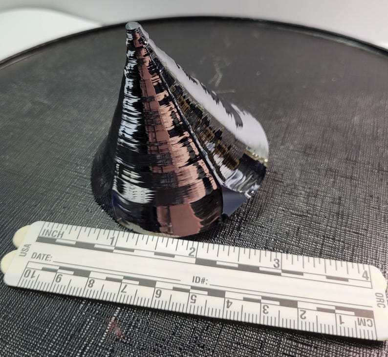 Puede incluir: Un objeto oscuro y reflectante en forma de cono con una base plana, posiblemente una oblea de semiconductor. Una regla blanca con marcas en cm est&aacute; en primer plano. La regla muestra medidas de hasta 10 cm.