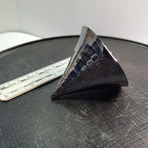 Puede incluir: Un objeto triangular oscuro y met&aacute;lico con una superficie reflectante. Una regla con marcas de cent&iacute;metros est&aacute; al lado del objeto. El objeto parece ser una pieza de un componente m&aacute;s grande, posiblemente tecnol&oacute;gico.