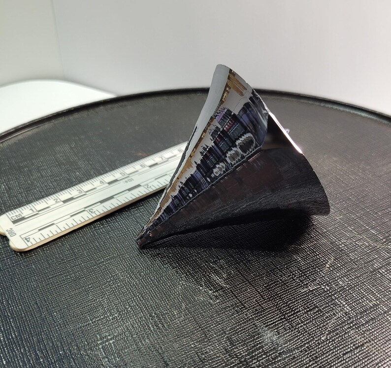 Puede incluir: Un objeto triangular oscuro y met&aacute;lico con una superficie reflectante. Una regla con marcas de cent&iacute;metros est&aacute; al lado del objeto. El objeto parece ser una pieza de un componente m&aacute;s grande, posiblemente tecnol&oacute;gico.