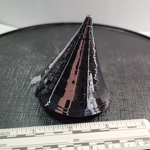 Puede incluir: Un objeto negro en forma de cono con una superficie reflectante, posiblemente de metal o material similar. El objeto est&aacute; colocado sobre una superficie oscura y texturizada. Una regla con medidas en cm est&aacute; en primer plano.