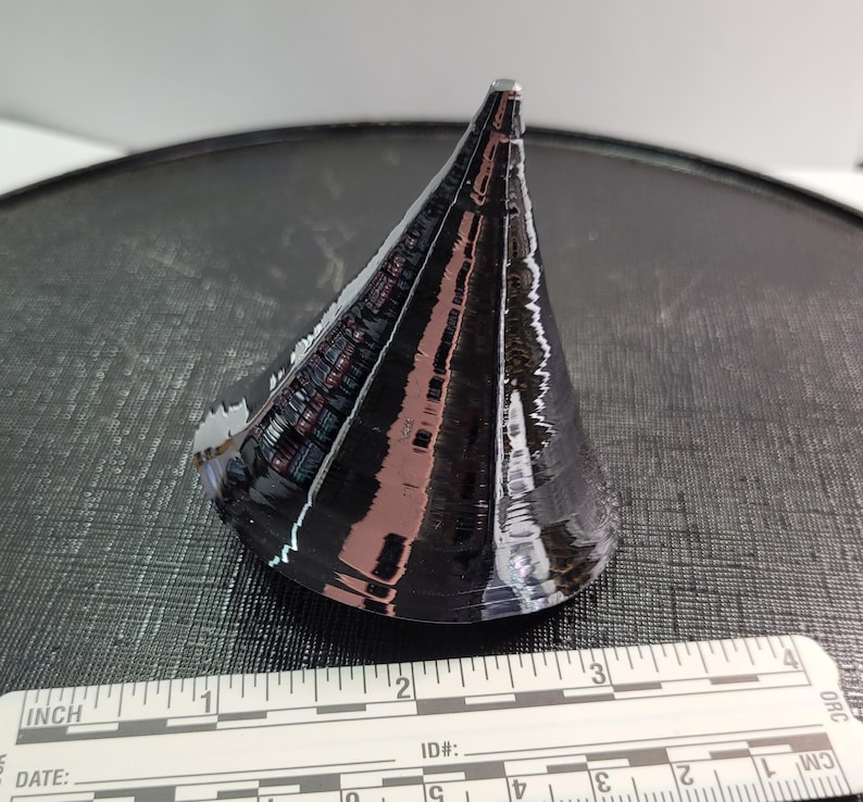Puede incluir: Un objeto negro en forma de cono con una superficie reflectante, posiblemente de metal o material similar. El objeto est&aacute; colocado sobre una superficie oscura y texturizada. Una regla con medidas en cm est&aacute; en primer plano.