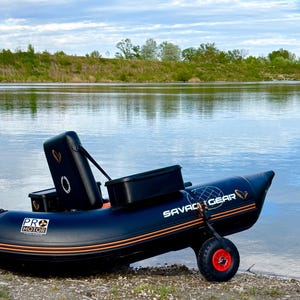 Peut inclure: Un bateau gonflable noir avec des accents orange et blancs, posé sur une rive de gravier. Le bateau porte les inscriptions "SAVAGE GEAR" et "PRO MOTOR". Une roue rouge est fixée au bateau.