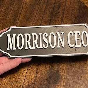 Könnte beinhalten: Ein schwarzes Holzschild mit weißer Beschriftung, auf dem "MORRISON CEO" steht. Das Schild hat einen abgerundeten, dekorativen Rand.