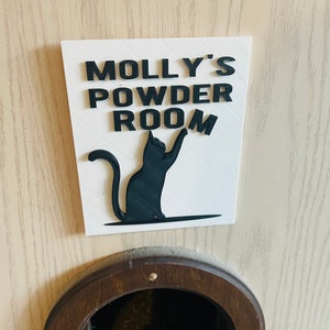 May include: White sign with black lettering that reads "Molly's Powder Room" with a black silhouette of a cat. The sign is mounted on a light wood surface with a brown cat door below.