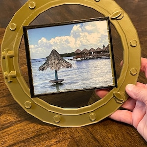 May include: A gold-colored, circular picture frame with a square photo of a tropical scene. The frame has a porthole design with decorative bolts and rivets. The photo shows a thatched-roof structure over the water.
