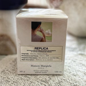 Puede incluir: Caja rectangular para una vela perfumada, con el texto "REPLICA" y "Maison Margiela PARIS". La caja tiene una pequeña imagen de una persona y enumera la fragancia como "Zen scents of matcha tea". Pesa 165 g.