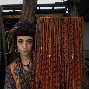 Puede incluir: Un conjunto de rastas sintéticas largas, rizadas, rojas y verdes. Las rastas están dispuestas en fila y cuelgan hacia abajo.