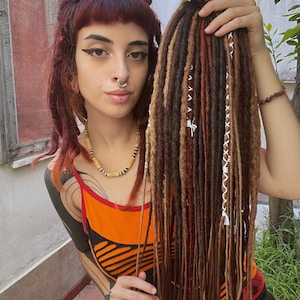 May include: A person with red hair and a nose ring is holding a set of long brown, blonde, and red dreadlocks with white string accents.