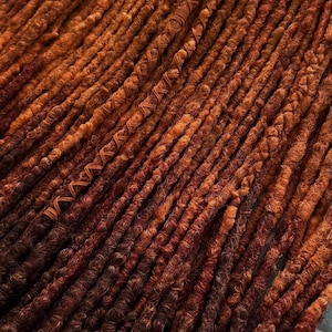 May include: Close-up of a bundle of brown and orange dreadlocks. The dreadlocks are tightly coiled and have a textured appearance. The color transitions from a lighter orange at the top to a darker brown at the bottom, creating a gradient effect. Some dreadlocks have decorative wraps.
