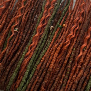 Könnte beinhalten: Nahaufnahme von handgefertigten Dreadlocks in verschiedenen Braun-, Orange- und Grüntönen. Einige Dreadlocks haben dekorative Perlen und Wicklungen. Die Dreadlocks sind diagonal angeordnet.