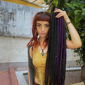 Può includere: Una persona con capelli rossi e dreadlocks neri e viola indossa una canotta gialla con dettagli in pizzo e una gonna marrone. Indossa un anello al naso argentato e una collana con un ciondolo di pietra verde.