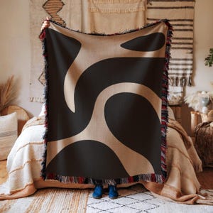 May include: A large, rectangular throw blanket with a black and beige abstract design. The blanket has a fringed edge with red, black, and blue accents. The blanket is held up in front of a bed with a neutral-toned bedspread.