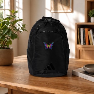 May include: Black backpack with a colorful butterfly design on the front. The backpack has side pockets and a large Adidas logo. The butterfly is embroidered with shades of blue, yellow, and purple.