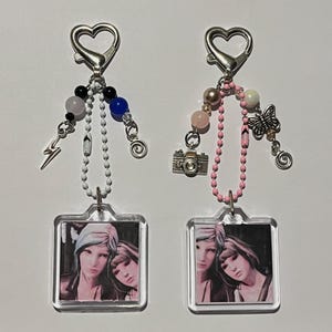 Può includere: Due portachiavi con chiusure a forma di cuore. Uno ha un ciondolo a forma di fulmine e una fila di perline nere, bianche e blu. L'altro ha un ciondolo a farfalla e una fila di perline rosa. Entrambi hanno un ciondolo quadrato con una foto di due donne.