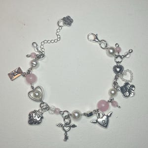 Pode incluir: Uma pulseira de berloques prateada com contas rosa, pérolas falsas e vários berloques. Os berloques incluem um morango, uma chave, um coração com asas e um pequeno envelope. A pulseira tem um fecho em forma de flor e um extensor de corrente.