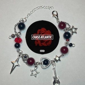 May include: A silver charm bracelet with red and black beads, a silver heart charm with a black eye, a silver star charm, a silver lightning bolt charm, and a silver guitar charm. The bracelet also has a silver clasp and a tag that says "CHASE ATLANTIC" with a parental advisory label.