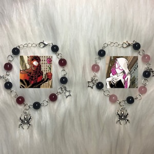 Könnte beinhalten: Zwei Armbänder mit Perlen mit Spider-Man- und Spider-Gwen-Comic-Kunst. Jedes Armband hat schwarze, rote und rosa Perlen, einen silbernen Spinnenanhänger und einen Sternanhänger. Die Armbänder sind durch eine silberne Kette verbunden.