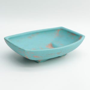 May include: A rectangular, turquoise ceramic bowl with a distressed finish. The bowl has a slightly curved shape and rests on three small feet. The turquoise color is accented with patches of a peachy-orange hue, giving it a rustic appearance.