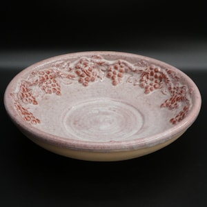 May include: A ceramic serving bowl with a light pink interior and a cream-colored exterior. The rim features a raised design of grape clusters in a darker pink hue. The bowl has a wide, shallow shape, suitable for serving.